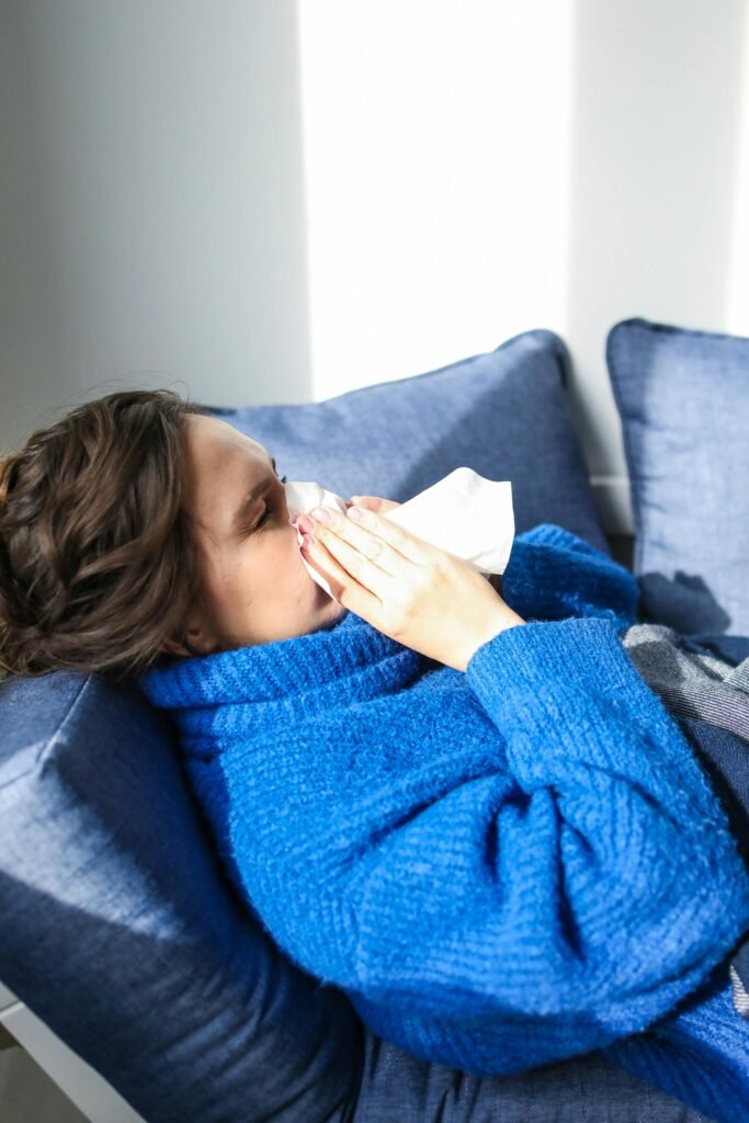 A woman in a blue sweater uses a tissue, lying indoors, suggesting illness or flu symptoms.